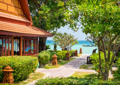 Tropischer Gartenweg mit Blick auf das türkisfarbene Meer, flankiert von einem traditionellen Holzhaus und blühenden Bäumen – Beispiel für hochwertige Hotelfotografie in Strandlage