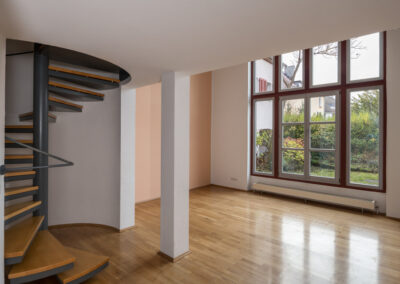 Lichtdurchflutetes Wohnzimmer mit glänzendem Holzparkett, einer freistehenden Spindeltreppe aus Metall und Holz sowie bodentiefen Sprossenfenstern, die den Blick in einen grünen Garten freigeben.