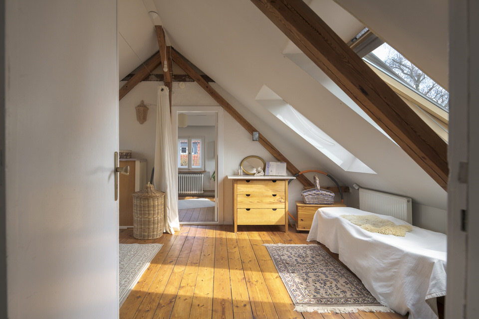 Authentic real estate photography of an attic room with wooden beams, wooden floor, single bed, chest of drawers and carpets under sloping ceilings with large windows