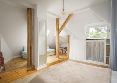 Helles, modern eingerichtetes Dachgeschoss-Schlafzimmer mit freigelegten Holzbalken, Echtholzboden, einem großen Spiegel und eingebauten weißen Schränken.