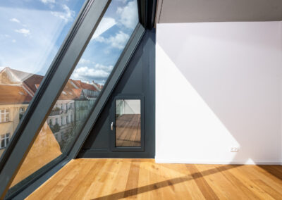 Ein moderner Raum mit hellen Holzböden, einem großen, schrägen Fenster mit Blick auf städtische Gebäude und einer kleinen Tür, die zu einem hölzernen Balkondeck führt.